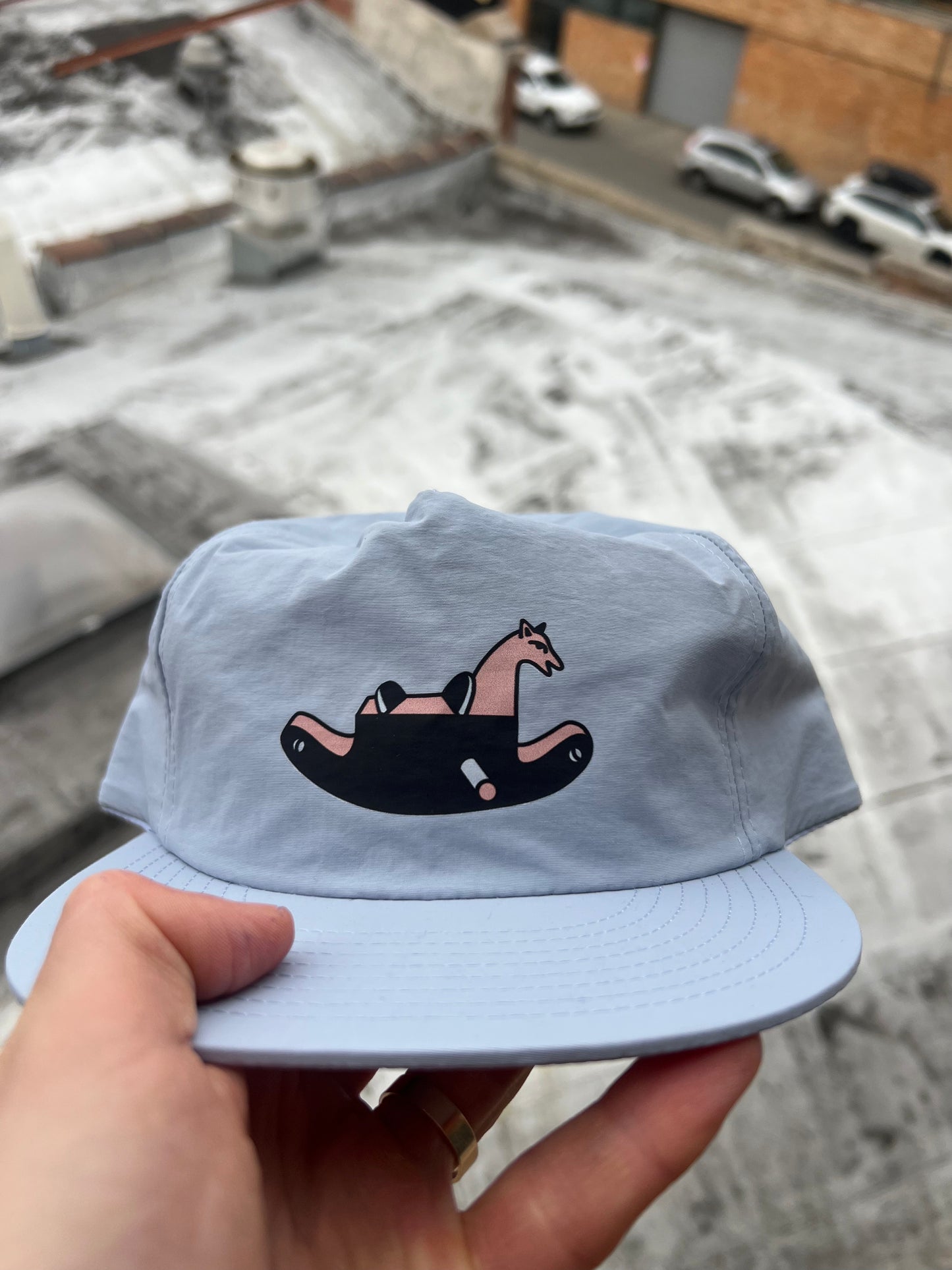 Light blue cap with a horse design held by a hand against a snowy ground.