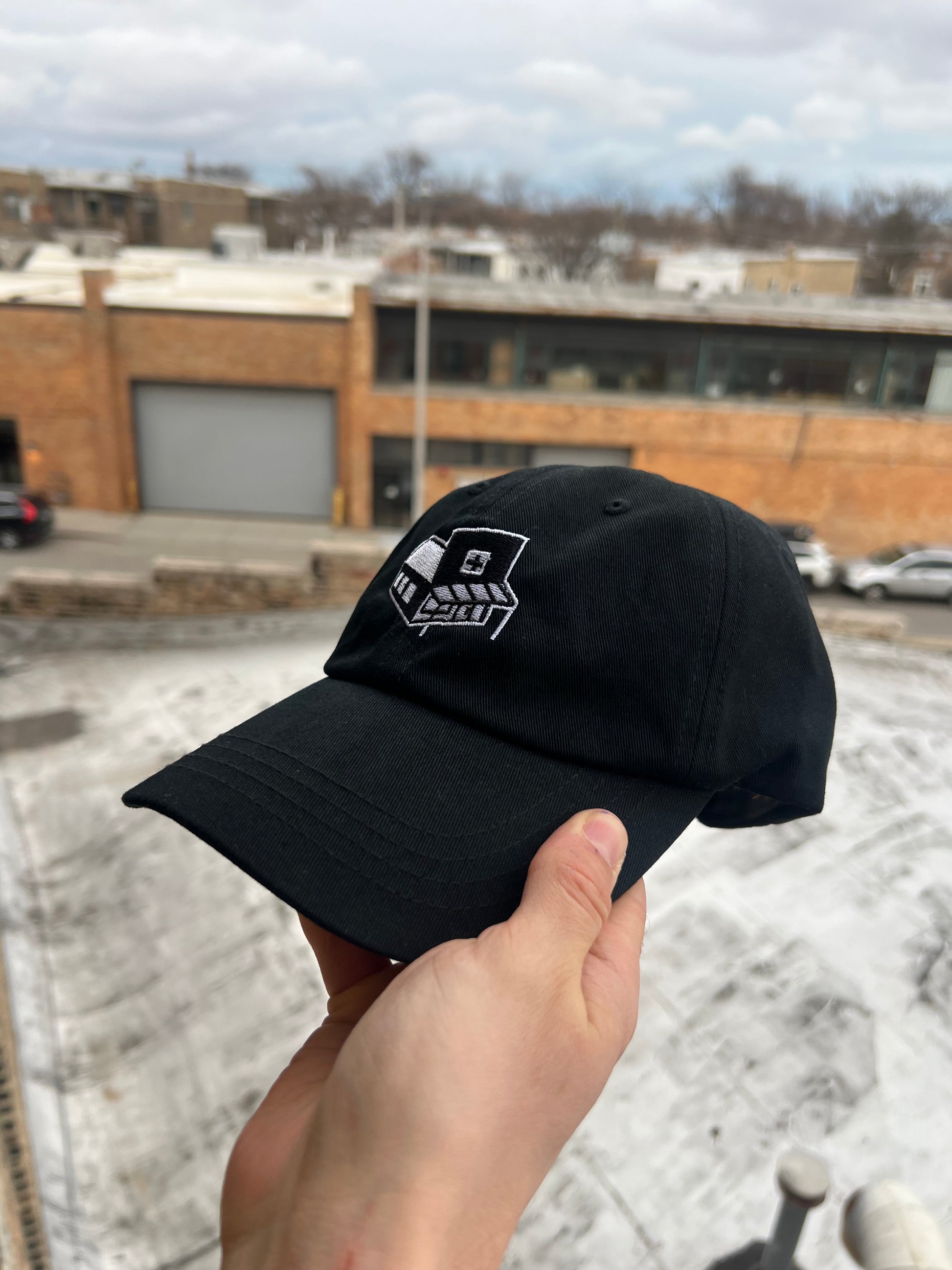 Black cap with a logo held by a hand on a rooftop with buildings in the background