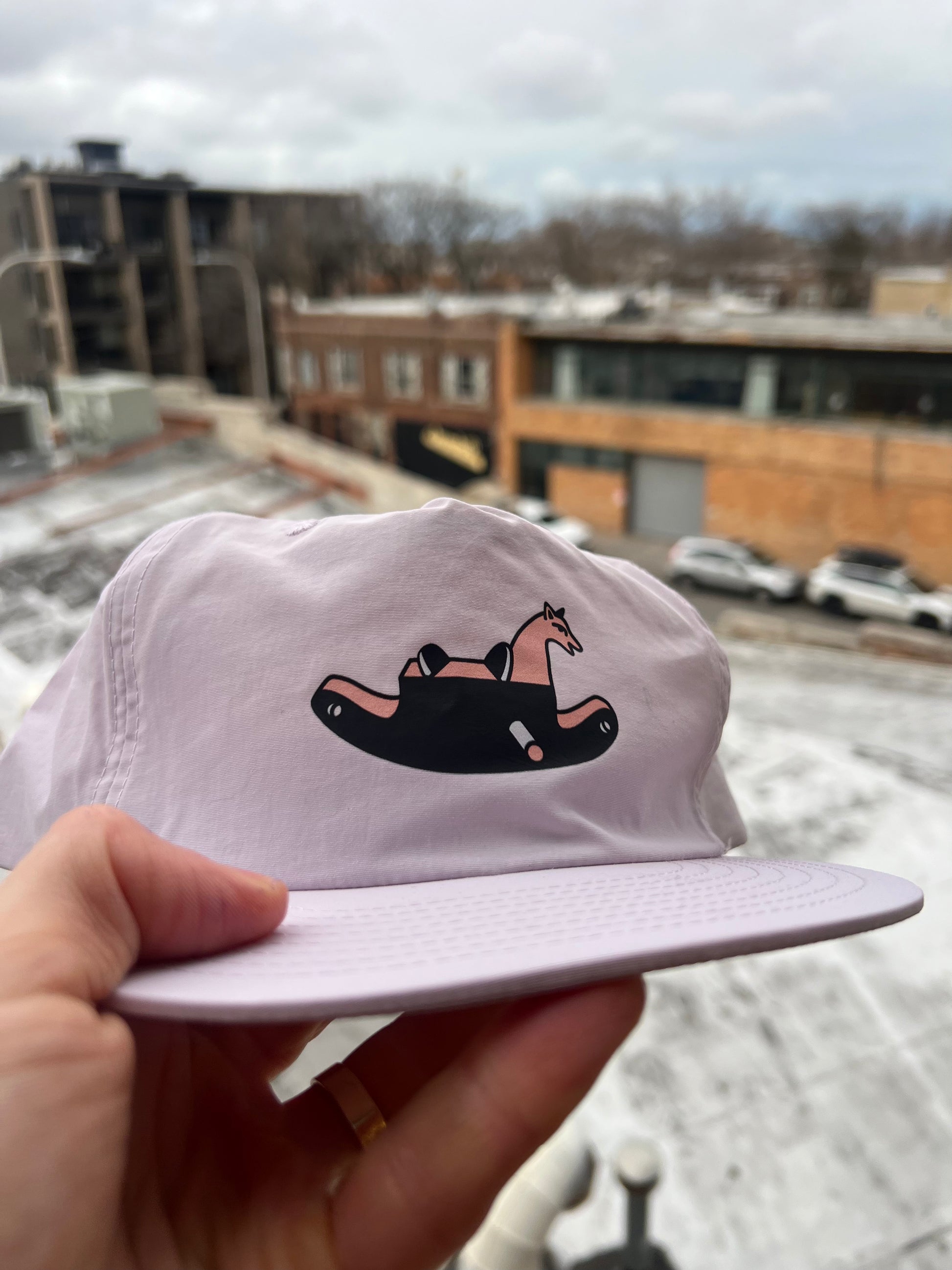 White cap with a black and pink design held in front of a snowy urban landscape.